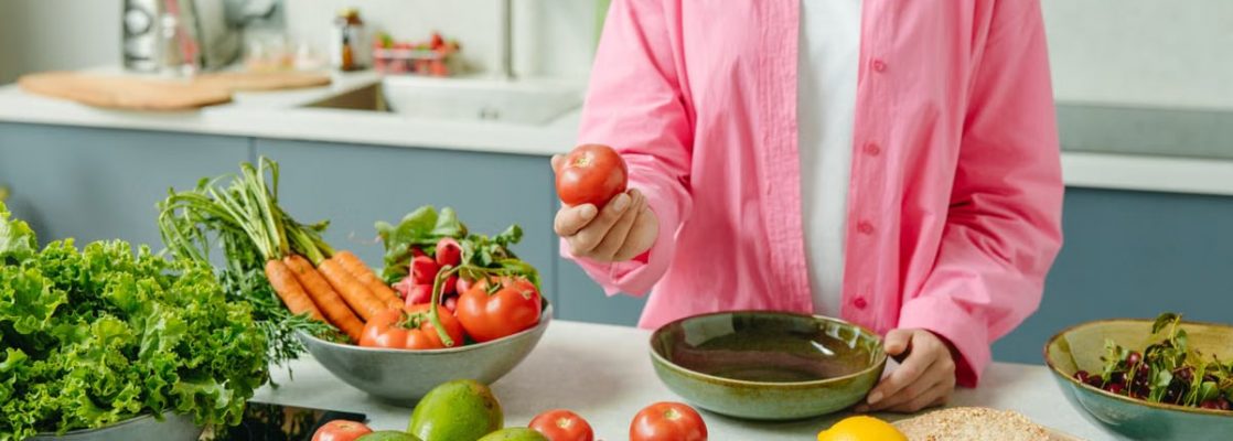 Woman in pink overshirt holds tomato in kitchen with fresh fruits and veggies.